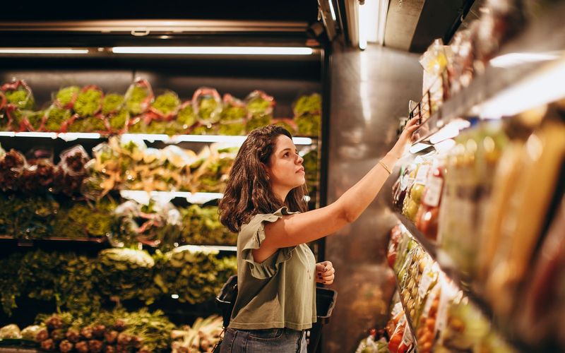 A woman selects fresh vegetables in a well-lit supermarket produce aisle